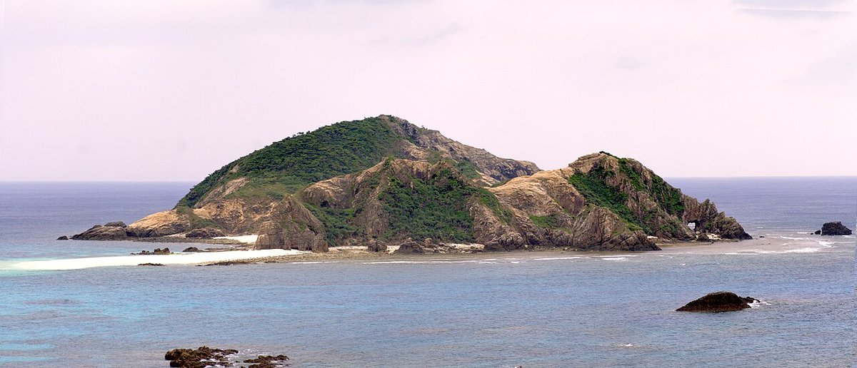 An uninhabited island seen from Aharen beach on Tokashiki, half green scrub and half bare rock, a white sandbar and pale green reef shallows in front of it, under a flat grey-white sky with no blue in it