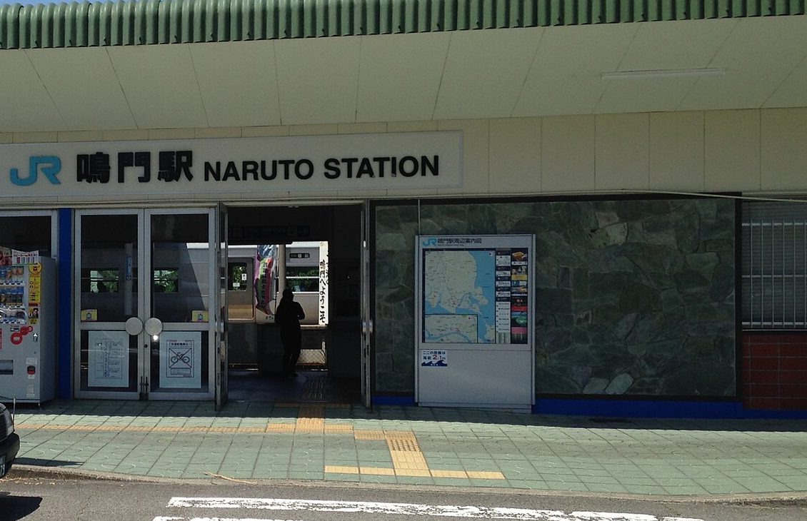 JR 鳴門站站房入口:白色招牌寫著「JR 鳴門駅 NARUTO STATION」,下方是玻璃門與通往月台的通道,可以看到停在月台的列車,右邊牆上掛著鳴門站周邊地圖看板