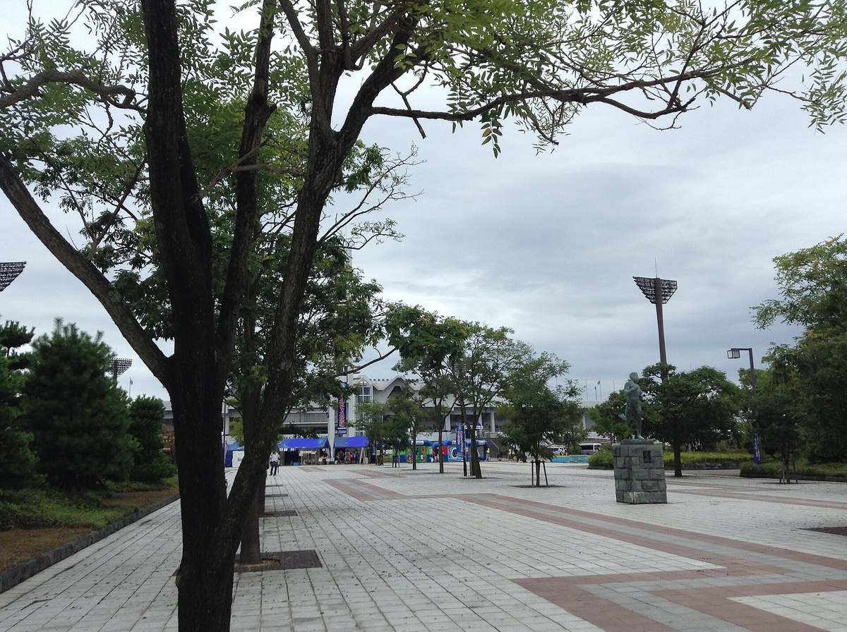 鳴門・大塚スポーツパーク園區內的寬闊地磚步道,兩旁種著行道樹,右邊石座上立著一尊跑者銅像,遠處是體育場的白色看台與照明燈塔,看台前搭著幾頂藍色帳篷