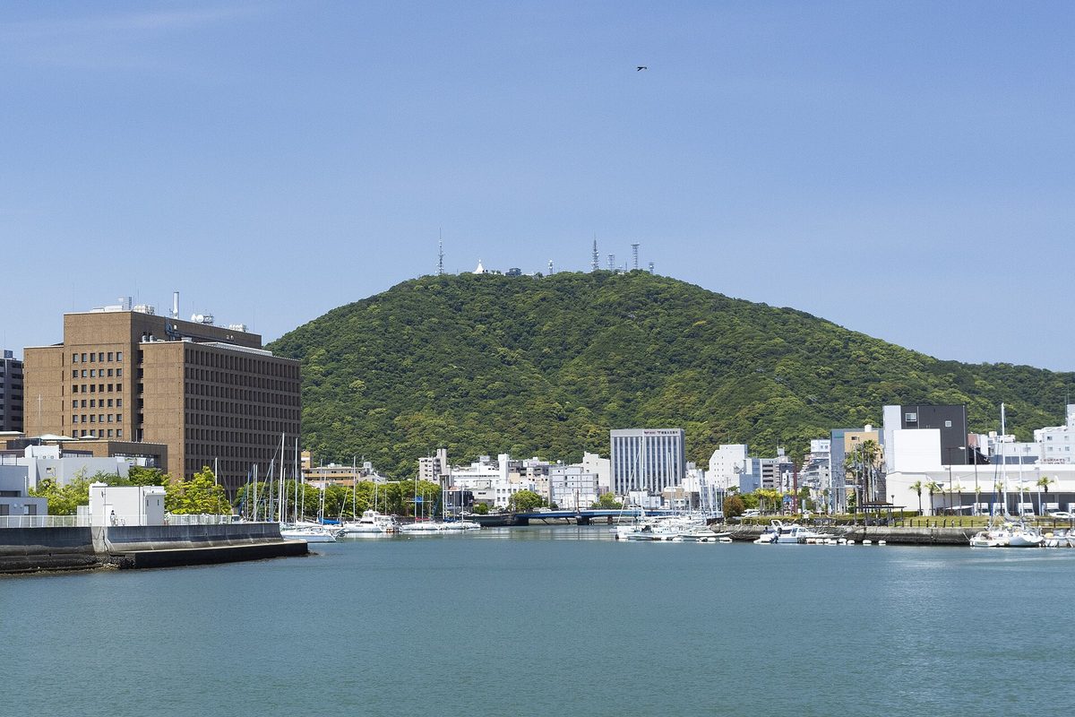 徳島市區的新町川:寬闊的河面上停著一排白色遊艇,兩岸是市區大樓,正後方是一座長滿綠樹的圓弧形山,山頂立著幾座電波塔