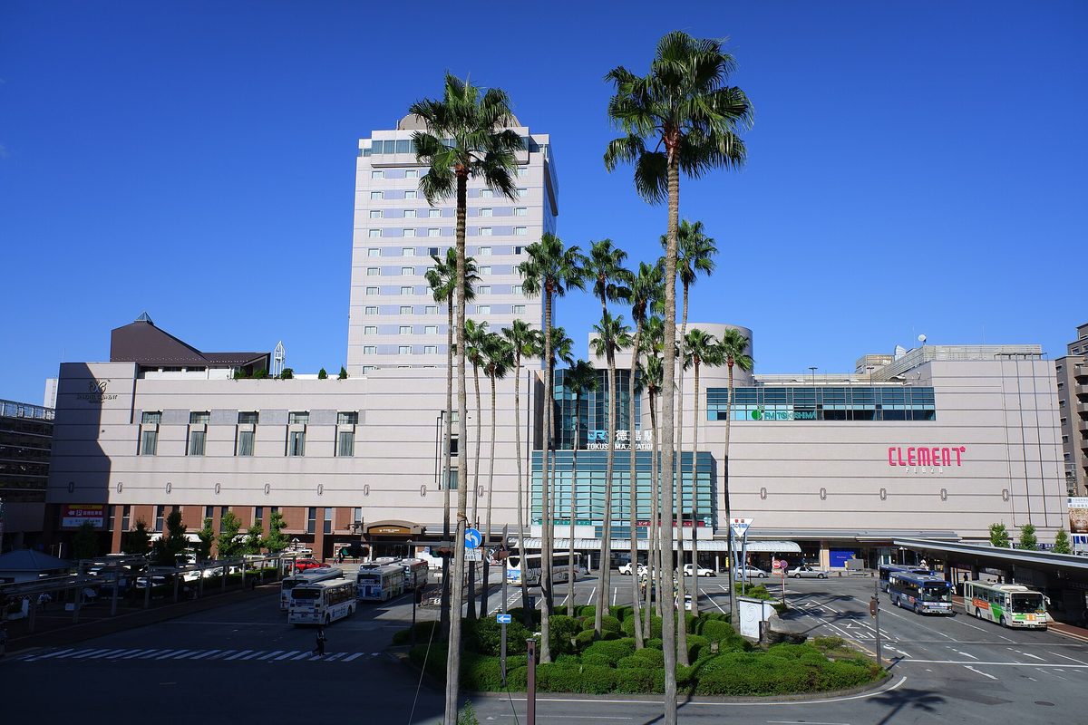 JR 徳島站站前:站房外牆寫著「JR 徳島駅 TOKUSHIMA STATION」,左後方是一棟高樓飯店,右邊是 CLEMENT PLAZA 商場,前方圓環種著一圈高大的棕櫚樹,周圍停著好幾輛路線巴士