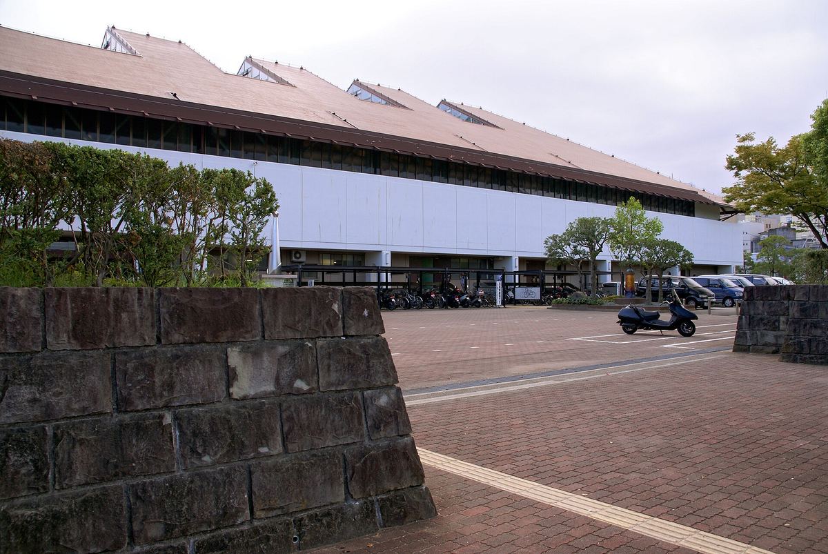 徳島市立體育館外觀:淺褐色的大斜屋頂上開著一排三角天窗,下方是長長的白色外牆,牆邊是腳踏車棚與停車場,前景是石砌圍牆與磚紅色地磚廣場