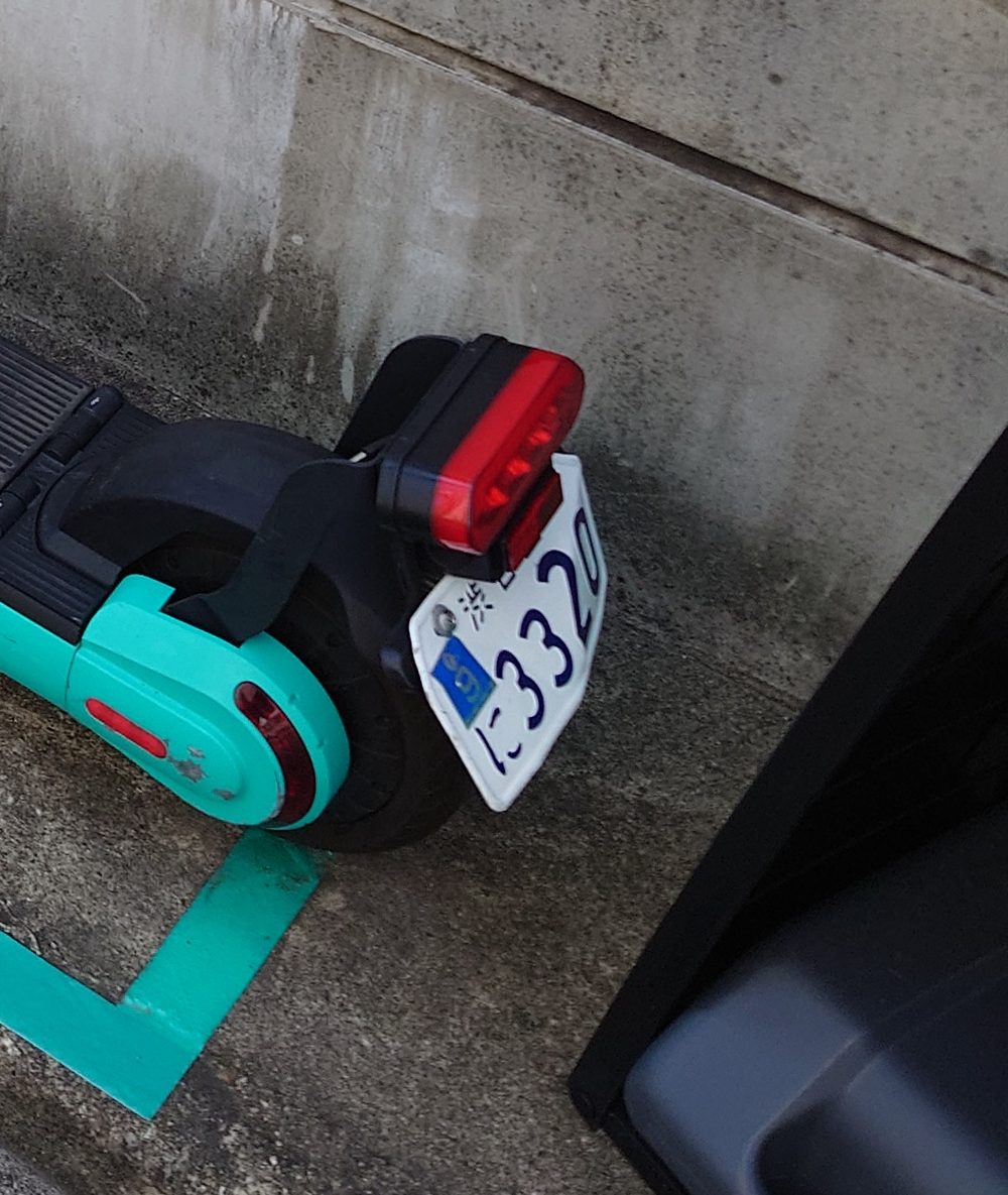 Close overhead view of the rear of an electric kick scooter: a horizontal red bar lamp sits above the black rear wheel, and below it hangs a small white number plate with the top and bottom corners cut to points, carrying blue characters reading Nagoya City and the number 3320, with a small blue sticker at its upper left; the turquoise rear mudguard and a red reflector strip are at the left, on concrete crossed by a turquoise parking line
