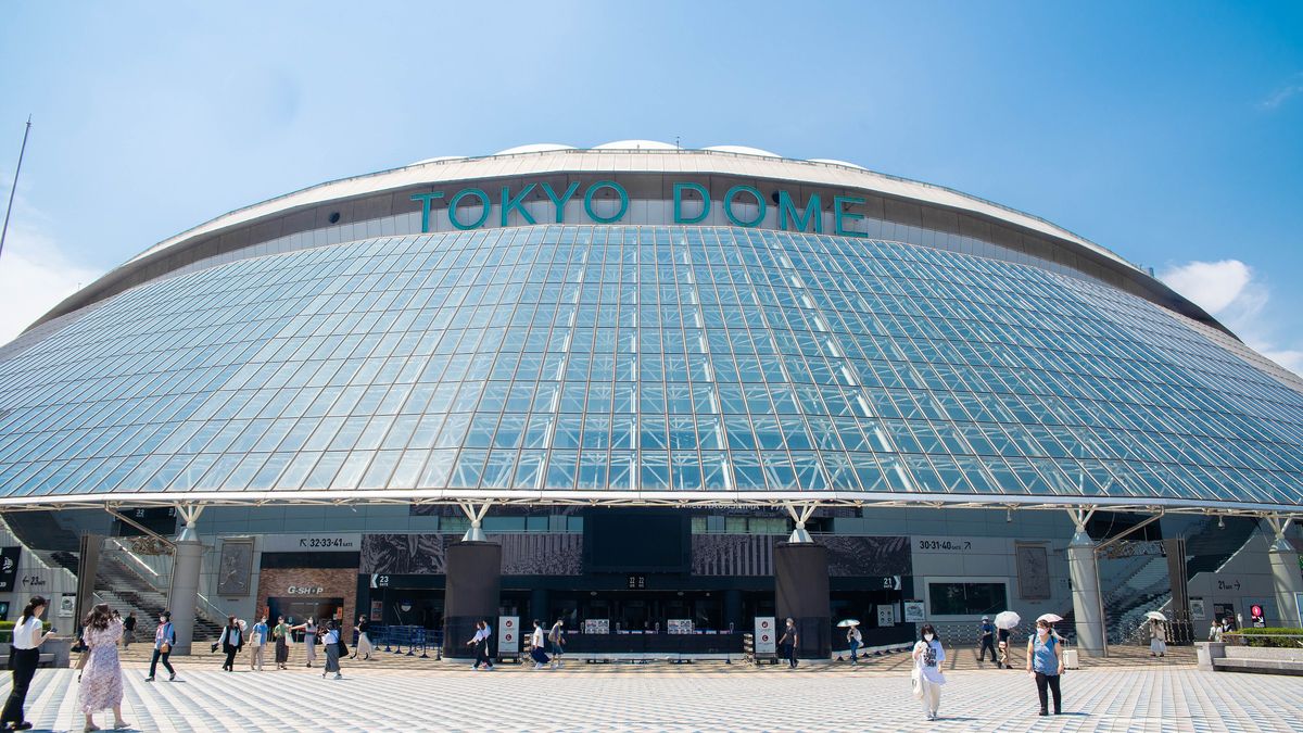 The exterior of Tokyo Dome, its white air-supported roof curving against a blue sky above signage and entrance gates, with pedestrians and trees on the plaza in front