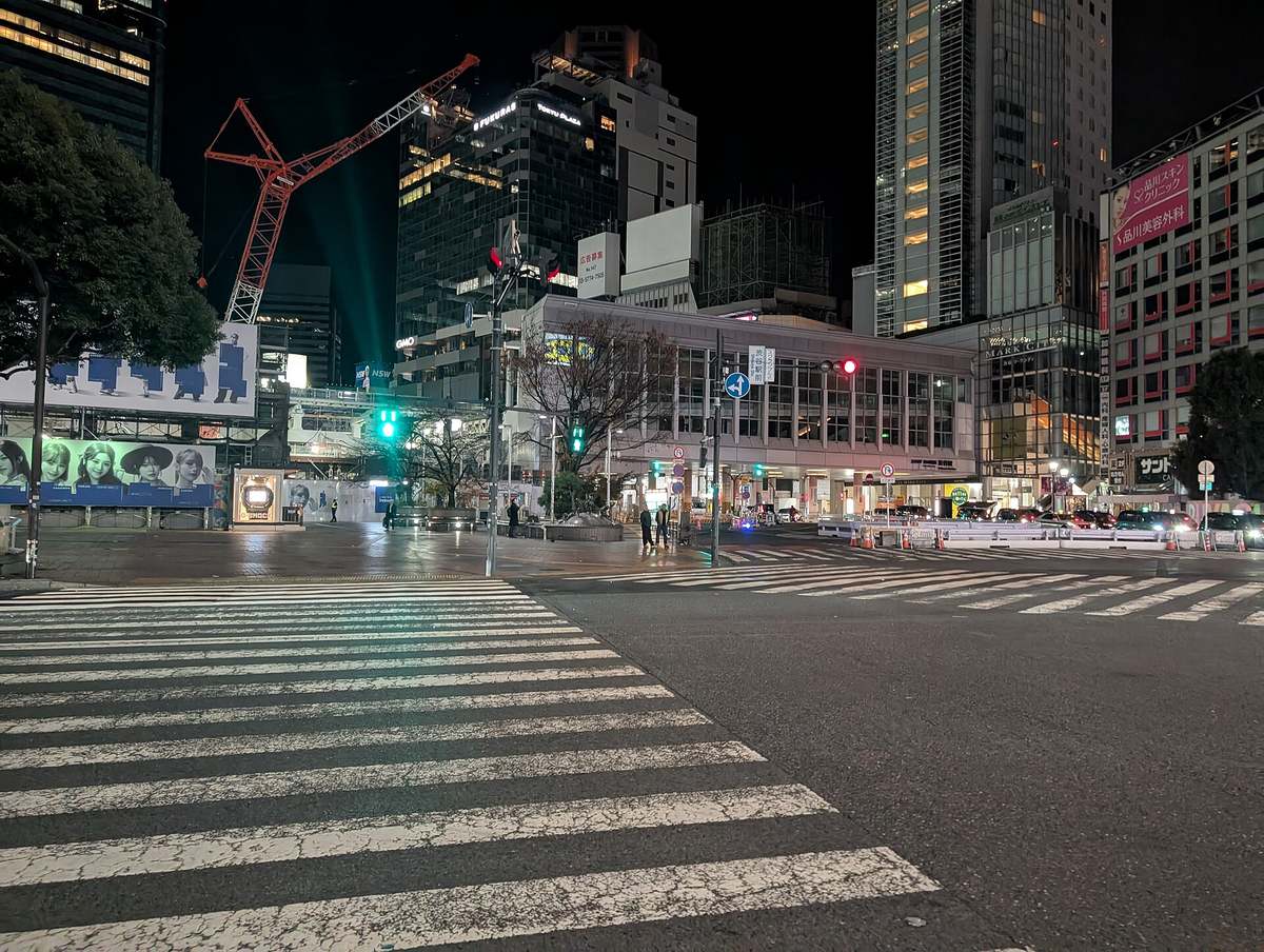 Shibuya's scramble crossing just before 2am on a weekday, the crossing and roadway all but empty apart from a few pedestrians, with a line of vehicles still waiting on the station side