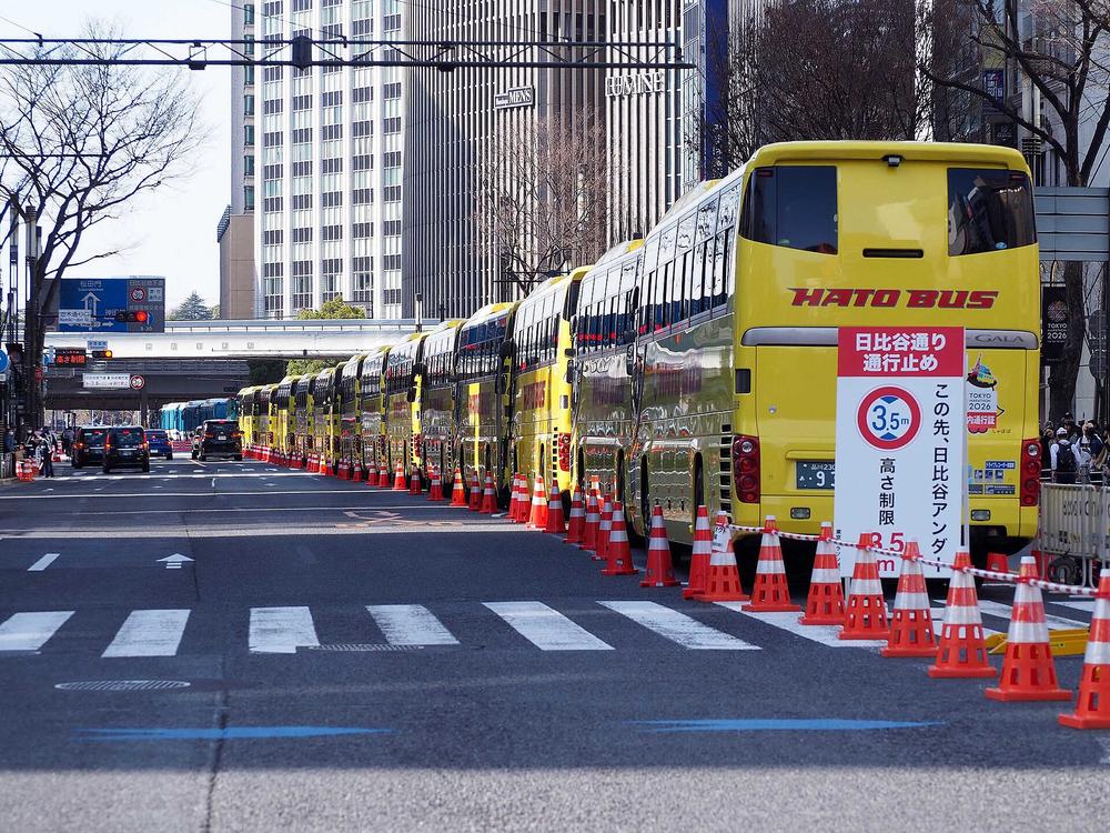 銀座晴海通的比賽日:一長排黃色はとバス停在道路中央、前面擺滿紅白三角錐,車尾立牌寫「日比谷通り通行止め」與高度限制 3.5m,左側車道仍有汽車行駛