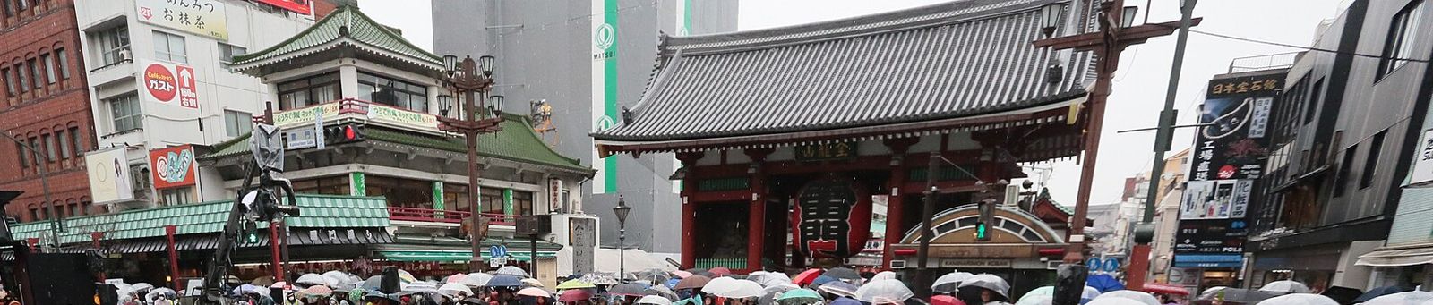 比賽日的淺草雷門前:大紅燈籠的雷門對面,沿著圍欄站滿撐傘的觀眾,左邊是搭著攝影機吊臂的轉角建築