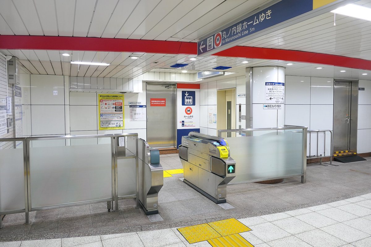 A lift-only ticket gate at Tokyo Metro Ogikubo station: one wide automatic gate showing a green arrow, a strip of yellow tactile paving running from the gate to a stainless lift door, a blue sign beside the lift reading ホームゆき to Platform, and a hanging sign overhead reading 丸ノ内線ホームゆき to Marunouchi Line 5m