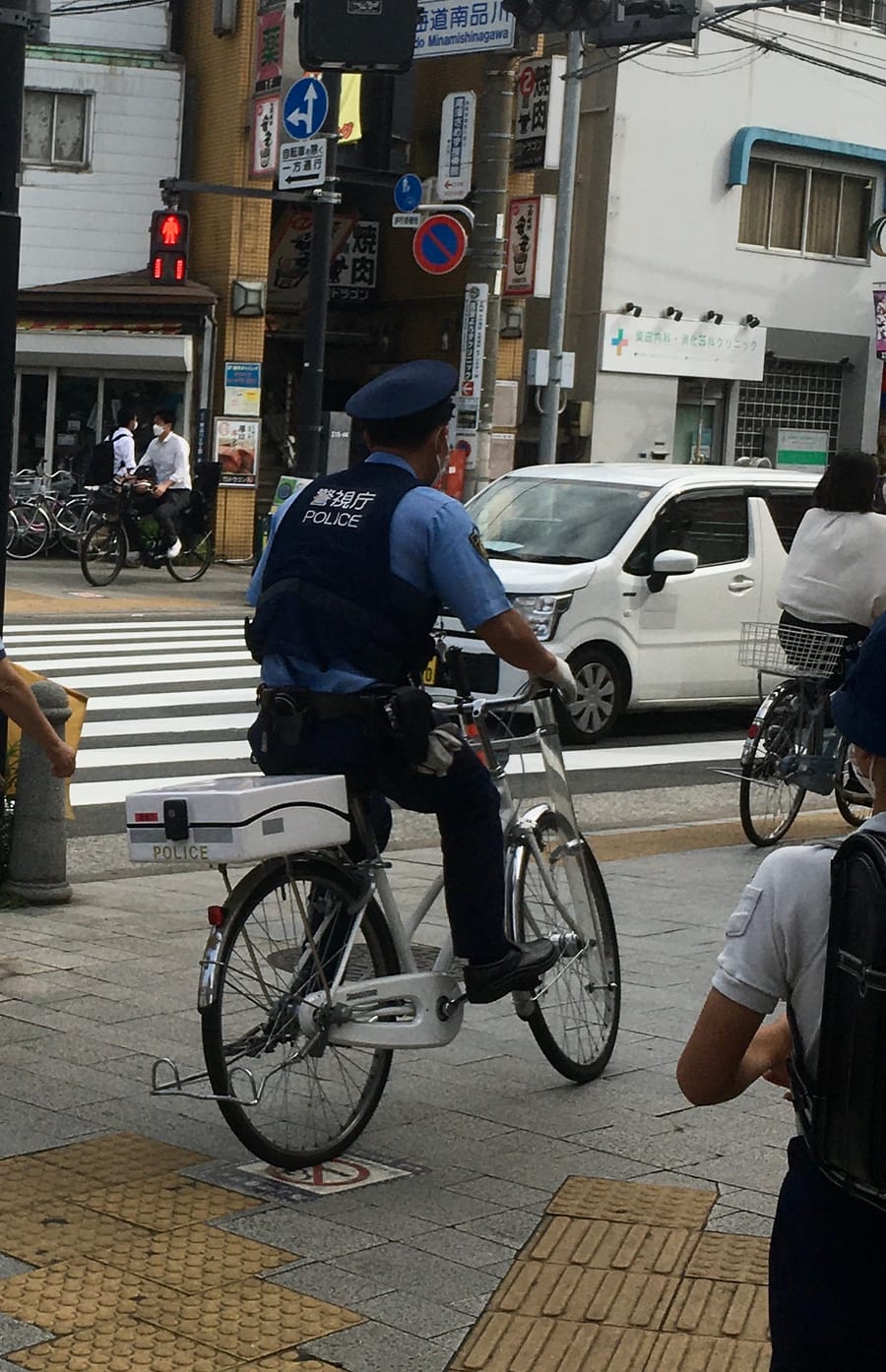 A Tokyo street corner: an officer in a pale blue uniform with 警視庁 POLICE printed across the back of his vest sits astride a white patrol bicycle waiting at a crossing, a white POLICE case mounted on the rear rack, the pedestrian signal opposite showing a red figure, and an ordinary cyclist standing alongside