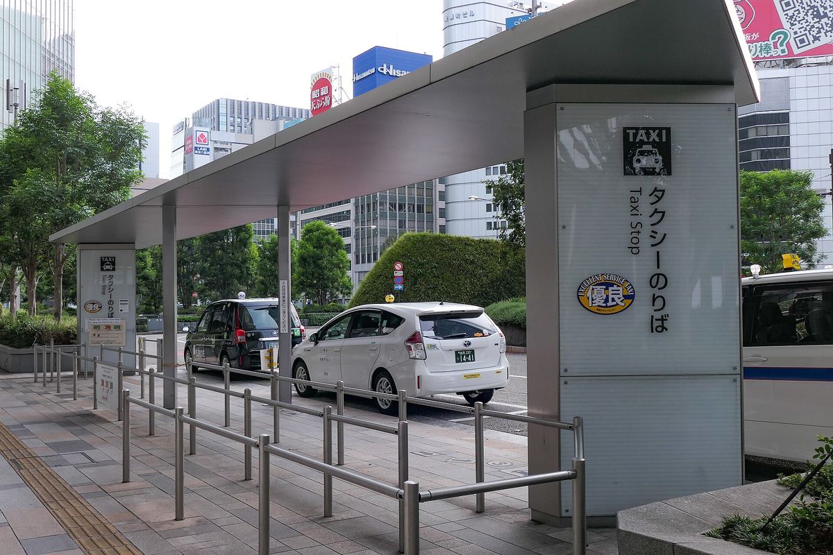 The taxi rank at the Yaesu exit of Tokyo Station, with a tall Taxi Stop pillar, queue railings and several taxis waiting at the kerb