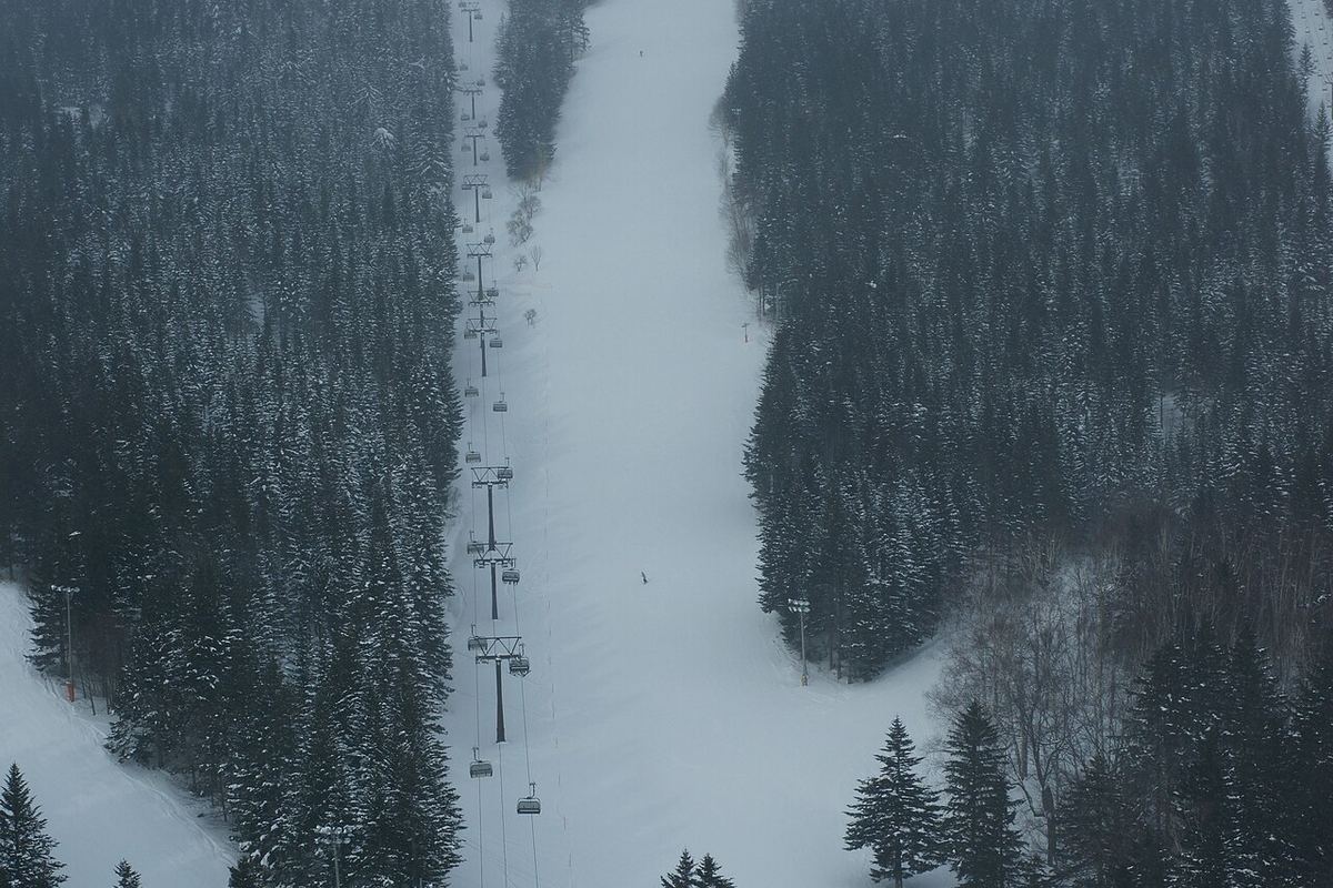 A wide run at Hoshino Resorts Tomamu dropping straight down the mountain, a line of chairlift chairs and towers climbing through the centre of the frame, snow-laden conifers on both sides and mist turning the summit grey-blue