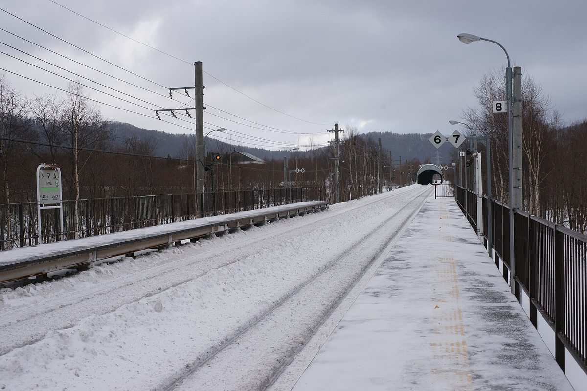 冬天的トマム駅 月台，路面積雪被踩實、鐵軌旁堆著除雪推起的雪堤，月台上空無一人，左側立著寫「トマム」的站名牌，前方軌道直直伸進一座隧道口，天空是厚重的灰雲