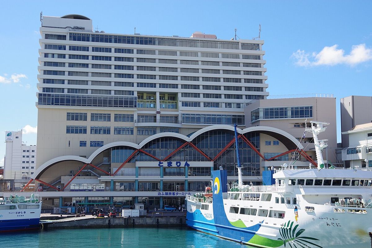 Tomarin, the passenger terminal at Tomari Port in Naha, three linked arched canopies over an entrance signed Tomari Wharf Passenger Terminal with a hotel block rising above it, the Tokashiki village ferry moored at left and an Aguni route ferry at right