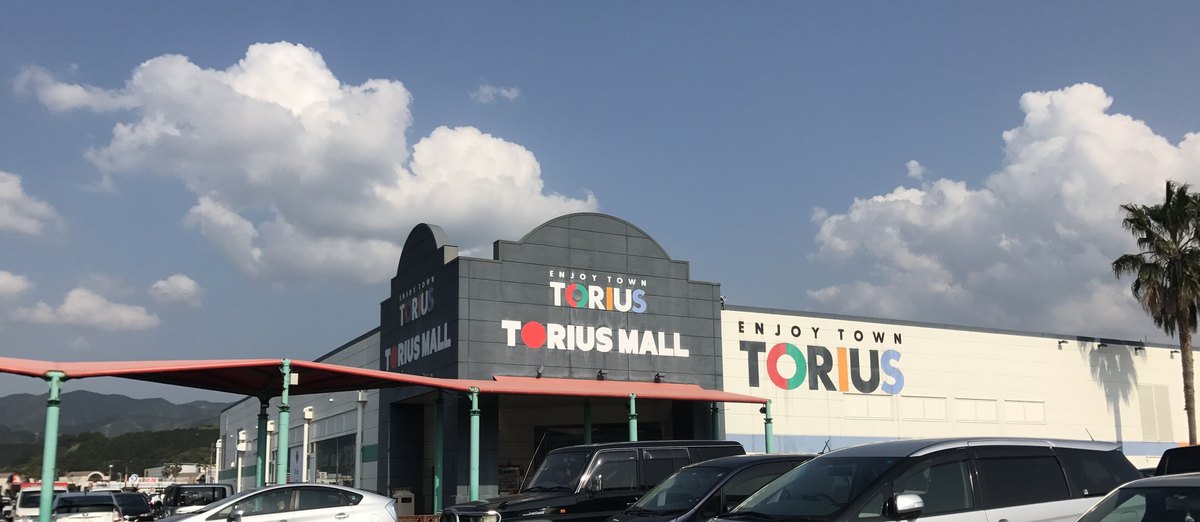 福岡縣久山町トリアス久山的商場外觀:深灰色門面上有 ENJOY TOWN TORIUS 與 TORIUS MALL 字樣,右邊白色外牆也有彩色 TORIUS 標誌,前面是停滿車的平面停車場,遠處有山