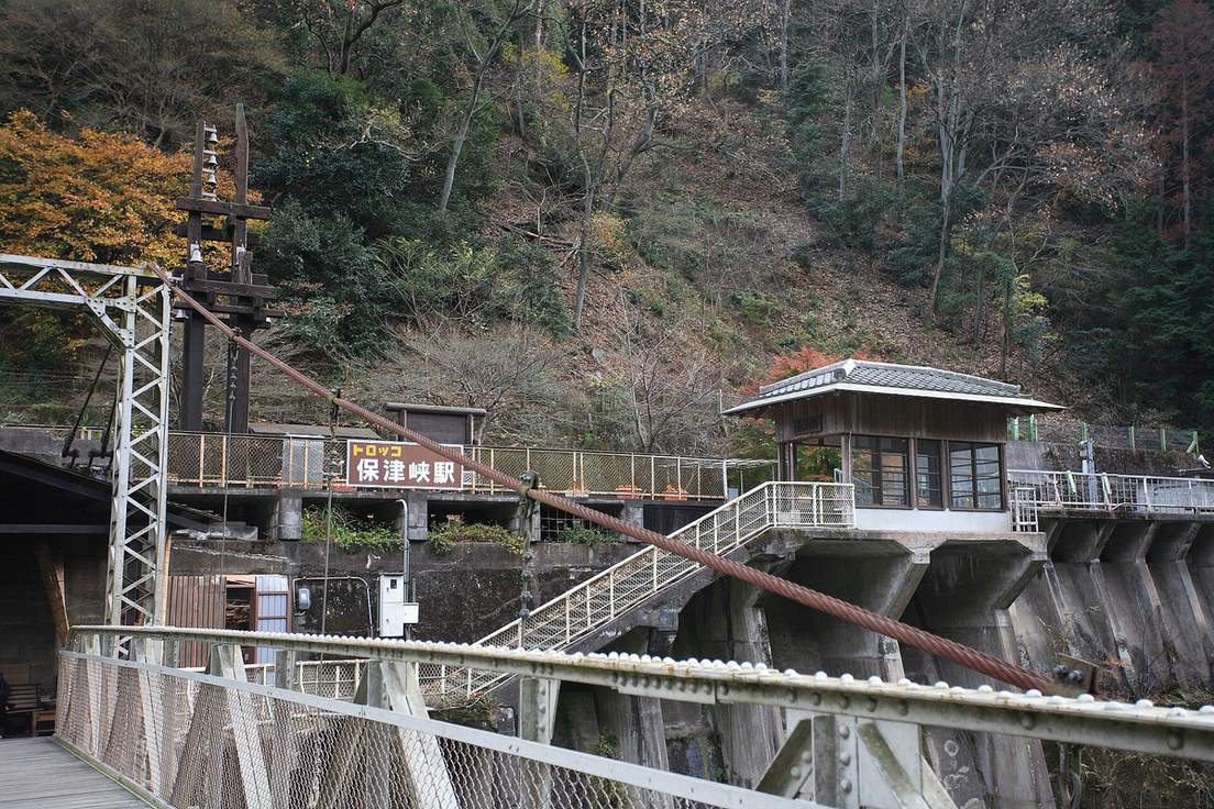 2006 年 12 月的小火車保津峽站：前景是吊橋的鋼索與欄杆，對岸混凝土高台上掛著「トロッコ保津峡駅」站牌，一道白色欄杆的樓梯從橋頭通往上方的月台，後方山坡有零星紅葉