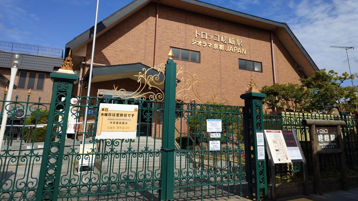 2026 年 5 月 20 日週三的小火車嵯峨站：綠色鑄鐵大門關著，門上貼著黃色告示「本日 水曜日は定休日です／Regularly closed on Wednesdays／今天是休息日」，後方紅磚色建築有「トロッコ嵯峨駅」金字，右側木牌寫「トロッコ嵯峨駅」