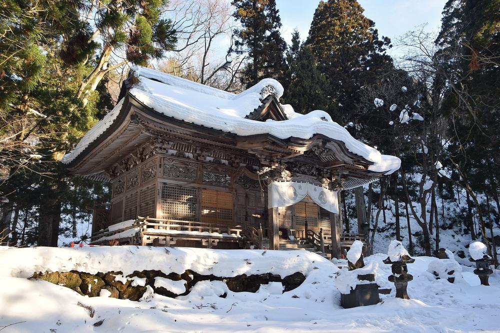 2022 年 2 月休屋的十和田神社拜殿：木造社殿的屋頂積著厚厚一層雪，簷下掛著白色的奉納布幕，前方的石垣與幾座石燈籠都頂著雪，四周是高大的杉樹