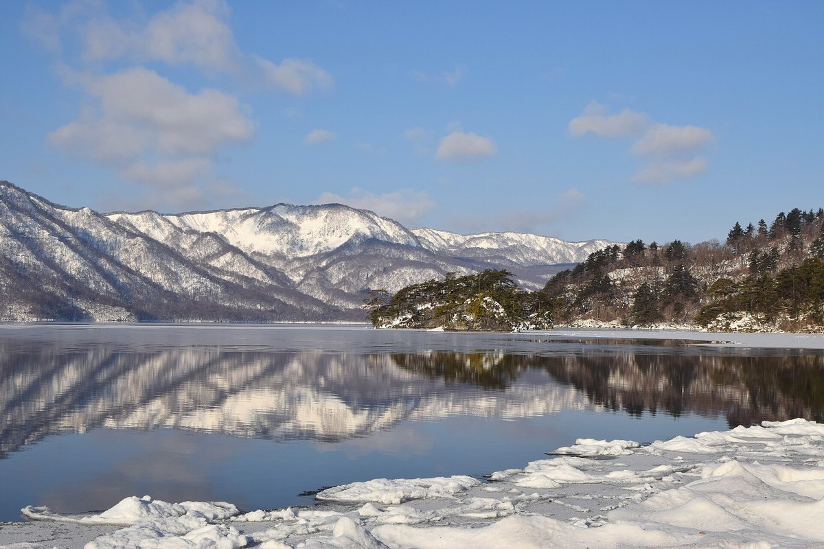 2022 年 2 月從休屋湖畔看十和田湖：近處岸邊是一塊塊碎裂的積雪與薄冰，湖面平靜、映著對岸覆雪的山，右邊是長著松樹的岬角，藍天有幾朵雲