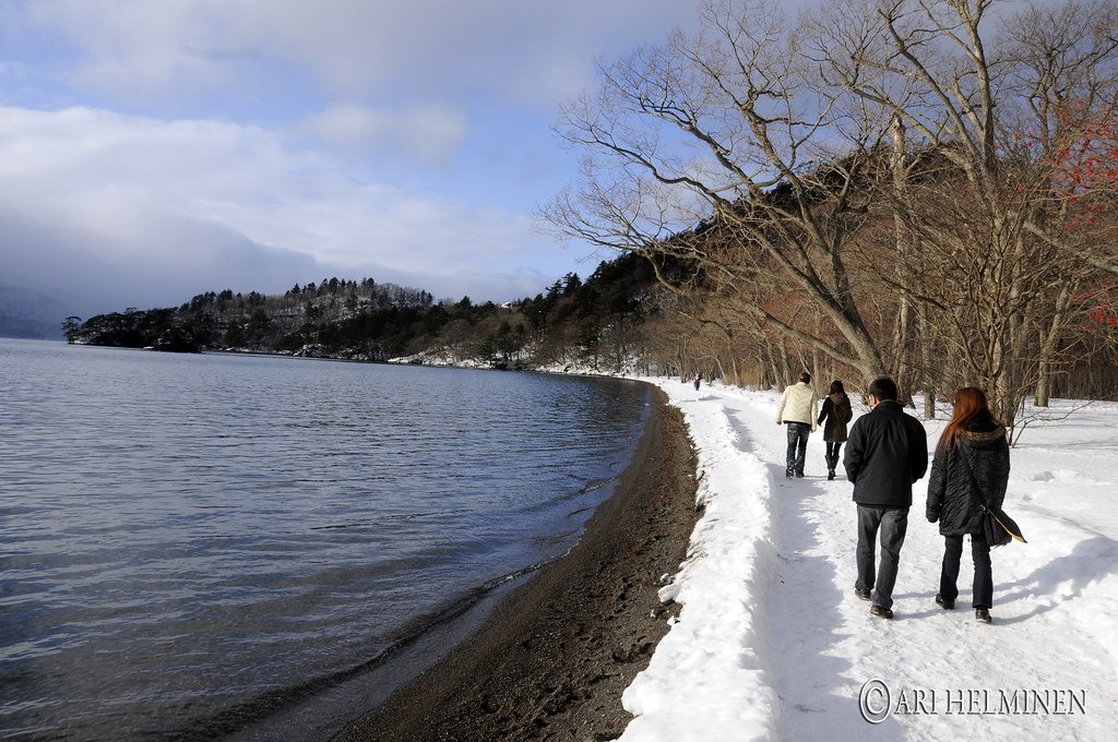 2012 年 1 月十和田湖畔的步道：左邊是湖水與深色的砂灘，右邊樹林下的步道整條被雪蓋住、中間踩出一條壓實的雪徑，四個穿冬季外套的人背對鏡頭往前走，遠處是長著樹的岬角，右下角有攝影者的浮水印