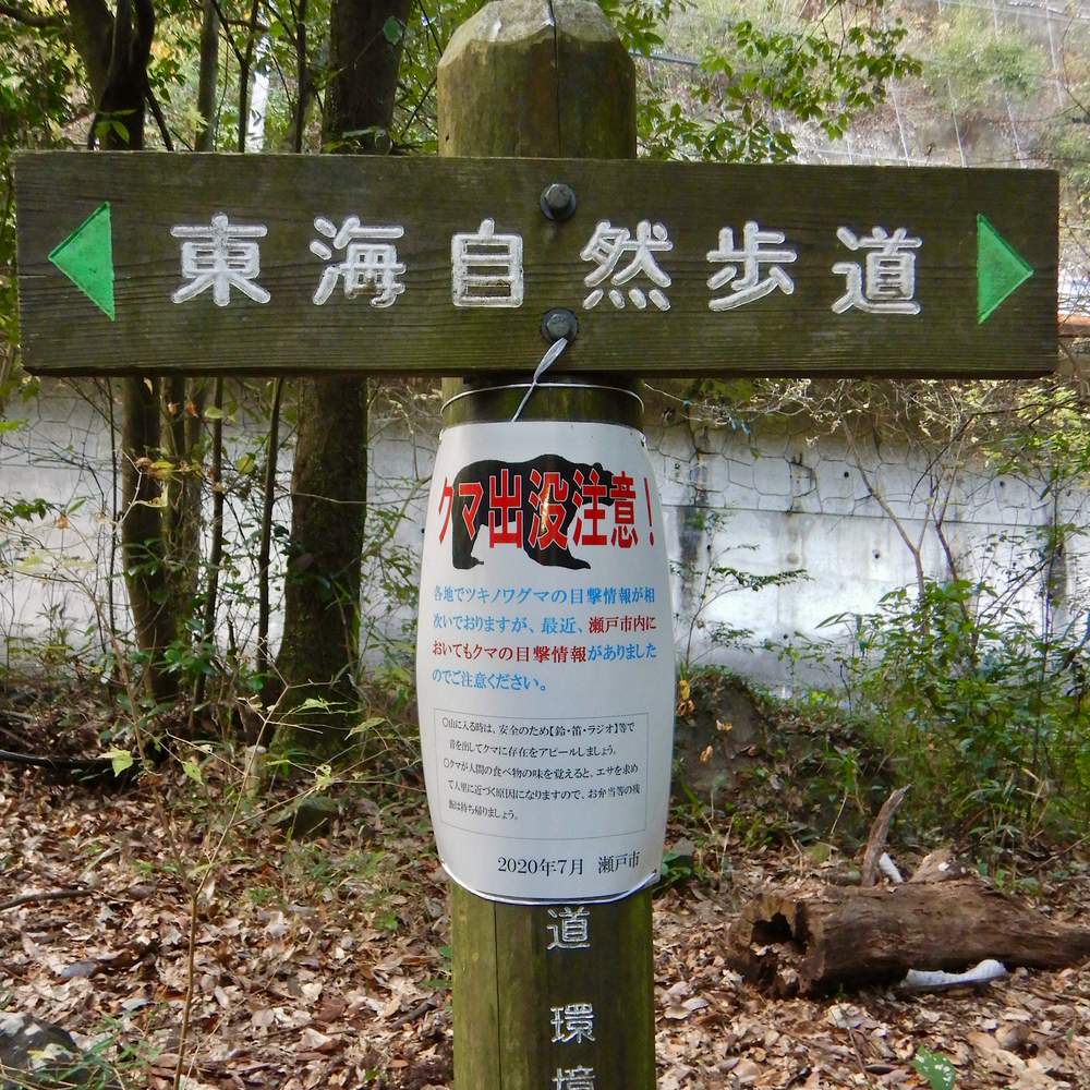 A wooden trail signpost on the Tokai Nature Trail in Seto City, Aichi, its crossbar carved with the trail name in Japanese between two green arrows, and a laminated bear warning notice tied to the post, dated July 2020 and issued by Seto City