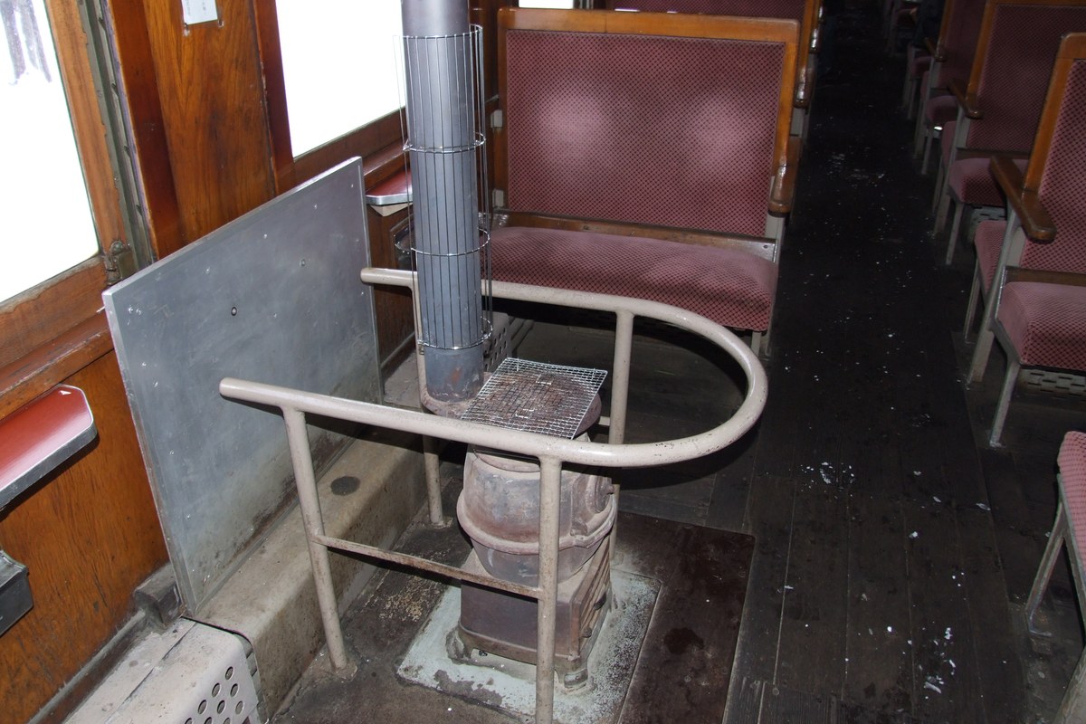 a cast-iron coal stove stands beside the aisle behind a guard rail with its flue running up to the ceiling, flanked by facing dark-red upholstered bench seats and a snow-white window