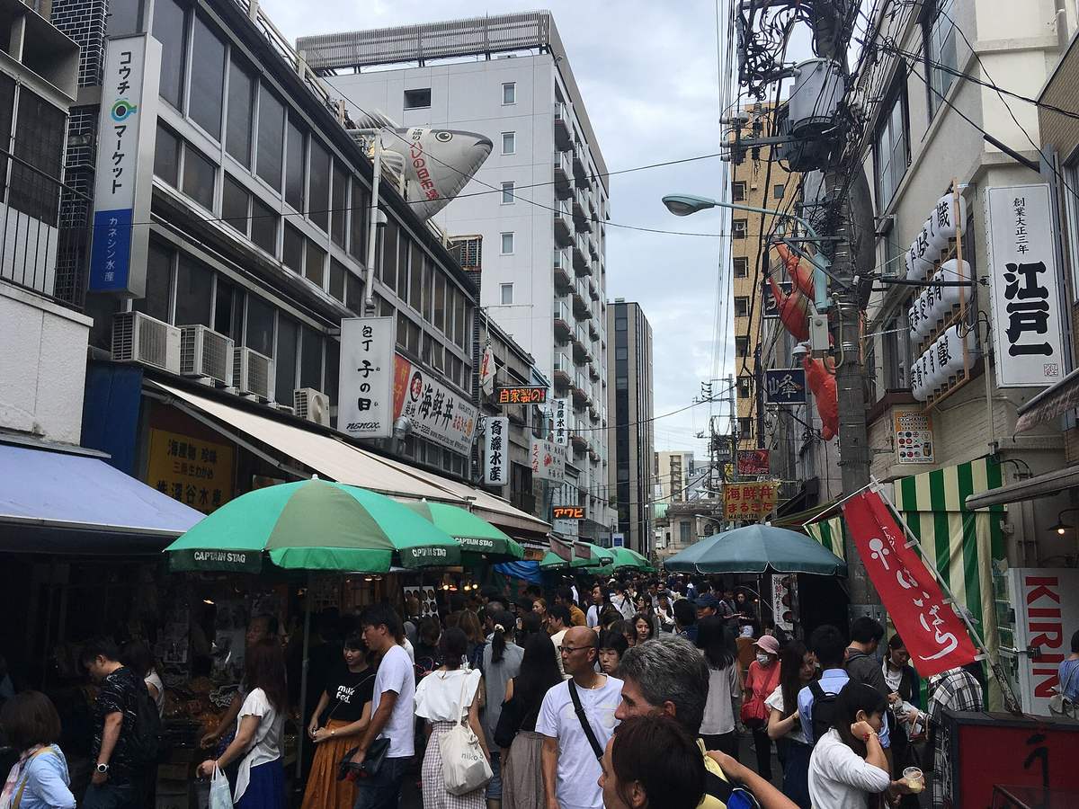 A lane in Tsukiji Outer Market packed shoulder to shoulder with visitors, seafood-bowl and dried-goods shopfronts under parasols and awnings running the length of the street