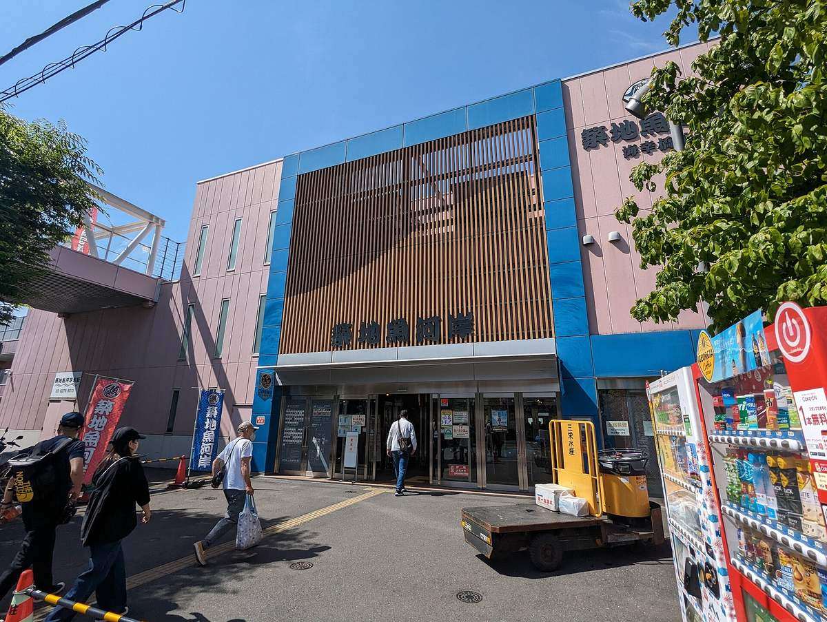 Exterior of the Tsukiji Uogashi Kaikobashi building, the Tsukiji Uogashi lettering set below a timber louvre screen above the automatic-door entrance, blue banners standing outside