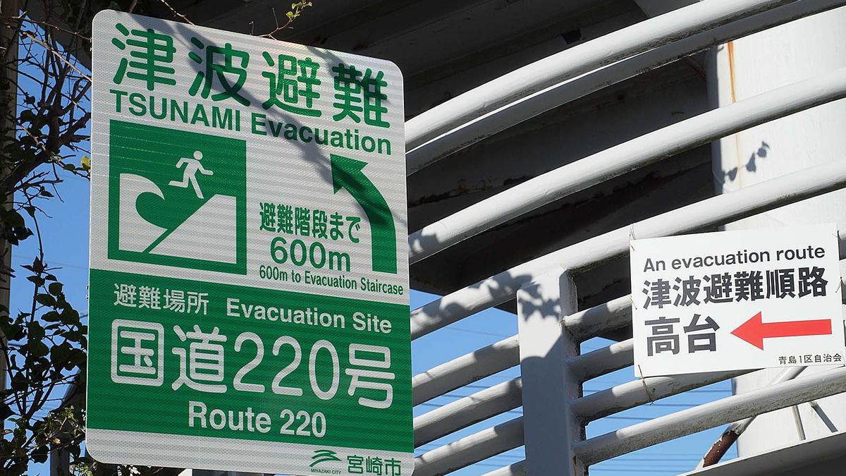 A tsunami evacuation route sign on the Japanese coast: green background with a white wave and running figure, and an arrow pointing toward higher ground