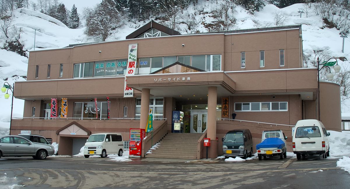冬天的 JR 津南駅站房：一棟兩層樓的淺棕色建築，正中的門廊上寫著「リバーサイド津南」，入口右側掛著直式木牌「津南駅」，二樓窗上貼著「温泉のある駅」的字樣，門口旁垂著一條白色直式布條。入口是一段水泥階梯，旁邊有紅色自動販賣機與紅色郵筒。站前廣場的柏油路面是濕的，路邊殘留剷過的雪，停著幾輛廂型車與輕型貨車；建物背後是一整片積雪的山坡與樹林，天空灰白