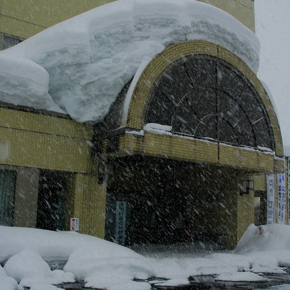 大雪紛飛中的津南町役場正門：米黃色磁磚外牆的建物，入口上方是一座半圓形拱窗，拱頂與旁邊屋簷上堆著厚厚一層積雪，積雪邊緣像一整塊白色厚板懸在屋簷外；入口左側牆上貼著一張紅字的「頭上注意」小告示，右側掛著幾條直式布條。前景是剷到路邊的雪堆與濕黑的地面，整個畫面佈滿斜斜落下的雪花