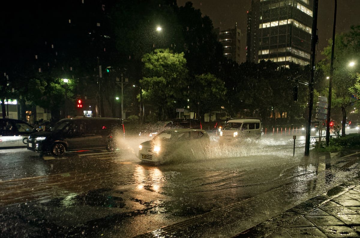 夜裡的強降雨:一輛車開過積水的十字路口,兩側濺起接近車身一半高的水花,柏油路面反著紅綠燈與車燈的光,空中看得到一條一條的雨絲