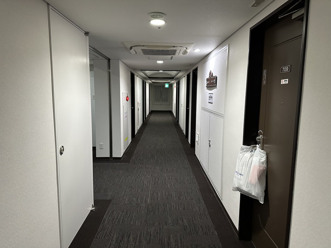 日本商務旅館的客房樓層走廊:兩側白牆與深色房門一路延伸到走廊底端,地面是灰色地毯,其中一扇門的門把上掛著洗衣袋,走廊上沒有人