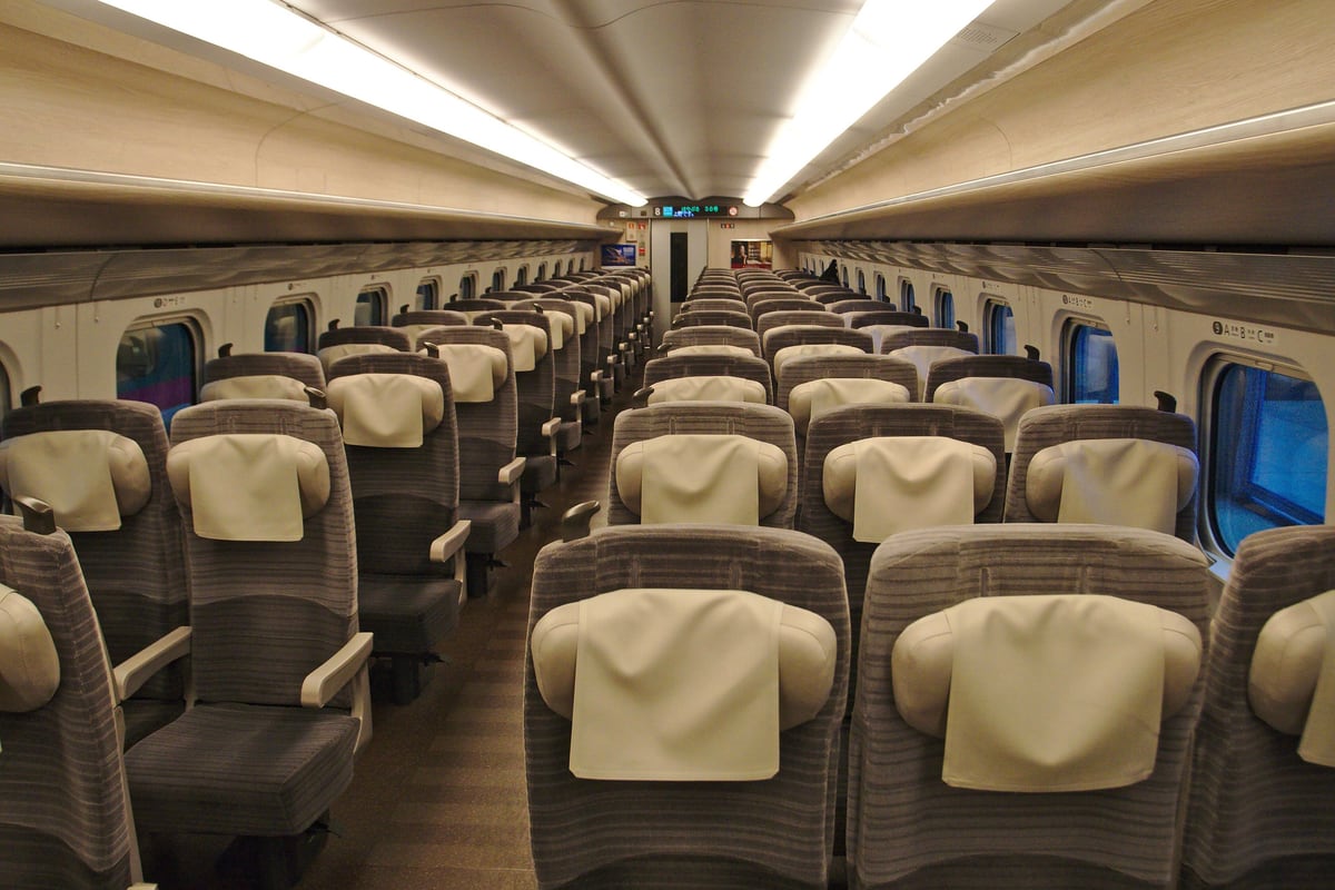 JR 東日本 E5 系新幹線普通車車廂內部,左側 2 人座、右側 3 人座,整排灰色椅背都罩著米白色頭枕布,走道空無一人;車廂盡頭的 LED 顯示器寫著 8 號車、はやぶさ 38 號與上野