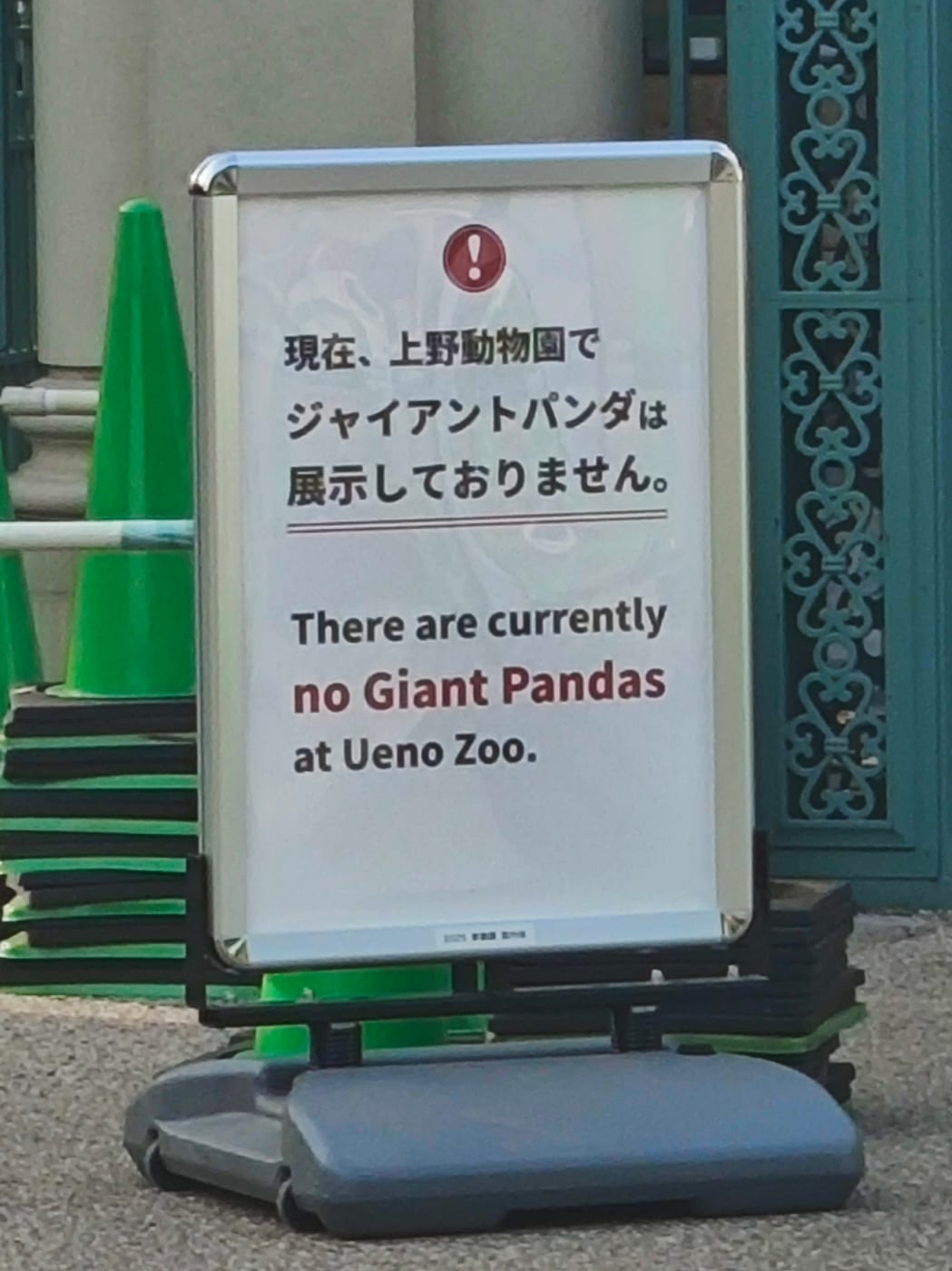 A silver-framed A-board outside Ueno Zoo. On a white panel under a red exclamation icon, Japanese text reads that giant pandas are not currently on display, with English below stating There are currently no Giant Pandas at Ueno Zoo. A teal wrought-iron gate stands to the right and green traffic cones are stacked behind