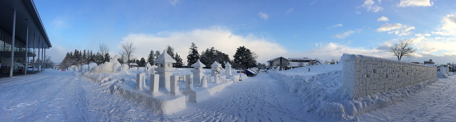 上杉雪燈籠祭會場白天的全景：左邊是玻璃帷幕、長屋簷的伝国の杜，中間一組沒點燈的雪燈籠方陣與幾座雪雕，右邊一道用雪砌成、刻出石塊紋路的雪牆，中央步道踩滿腳印，藍天白雲