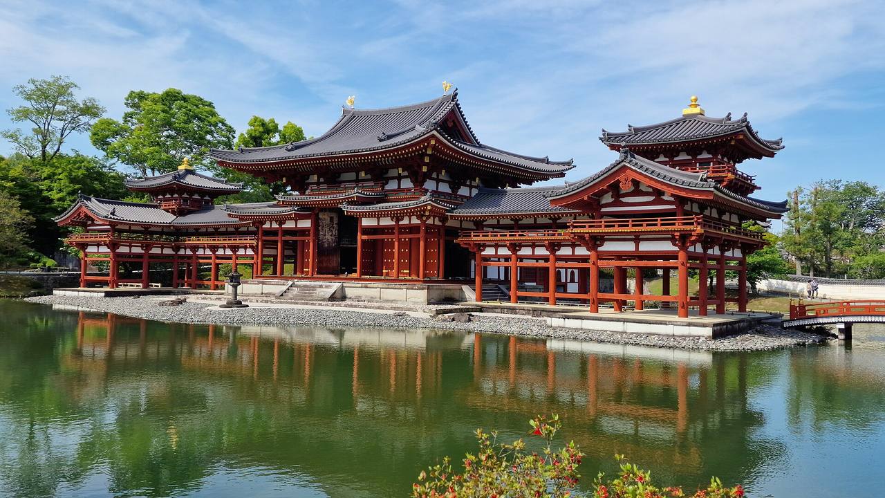 Byodō-in's Phoenix Hall in Uji seen at an angle across the Aji Pond on a clear day: vermilion central hall and wing corridors reflected in green water, a gilded phoenix at each end of the main roof ridge