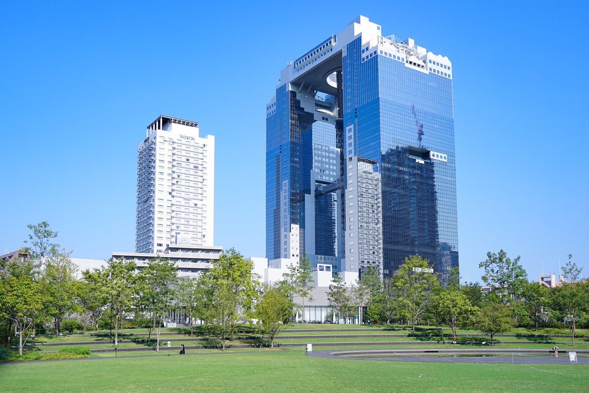 2025 年 8 月晴天,從大阪站北側的公園草地望向梅田藍天大廈:兩棟藍色玻璃帷幕塔樓在頂部相連、中間鏤空,左邊是白色的 WESTIN 飯店,前景是草地與一排樹