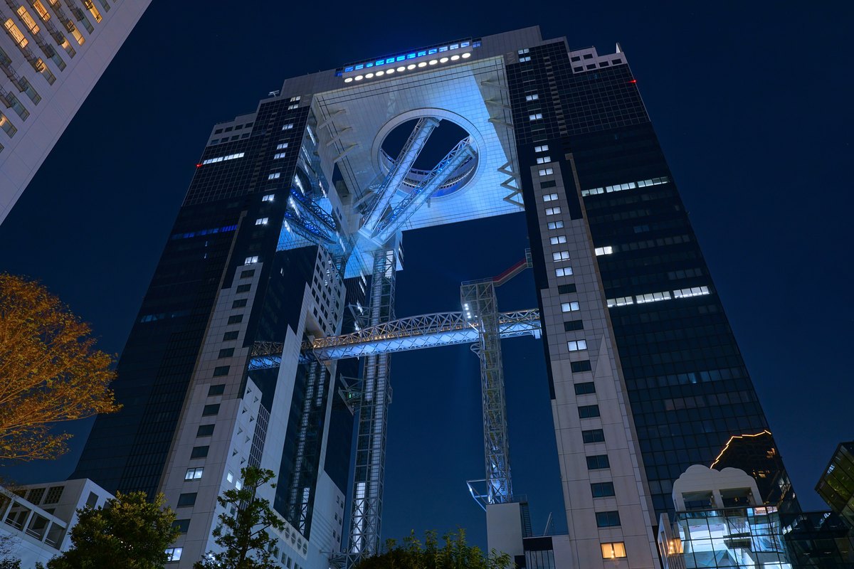 2025 年 8 月夜晚從地面仰望梅田藍天大廈:兩棟塔樓在頂部相連,連接處中央有一個打著藍白光的圓形開口,兩條斜向的手扶梯管穿過圓洞,下方另有一座空橋橫跨兩棟之間