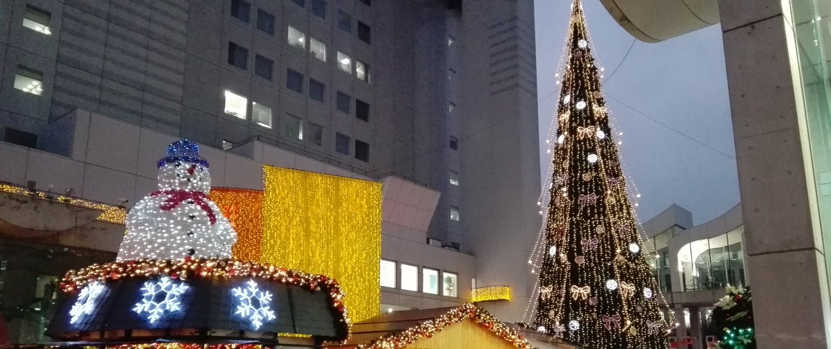 2015 年 12 月傍晚的梅田藍天大廈 1 樓廣場:右側是掛滿白色燈串與蝴蝶結燈飾的大型聖誕樹,左前方是屋頂站著發光雪人、屋簷有雪花燈飾的市集木屋,中間露出另一間木屋掛著彩球花圈的山形屋頂,後方牆面垂著金黃色燈簾