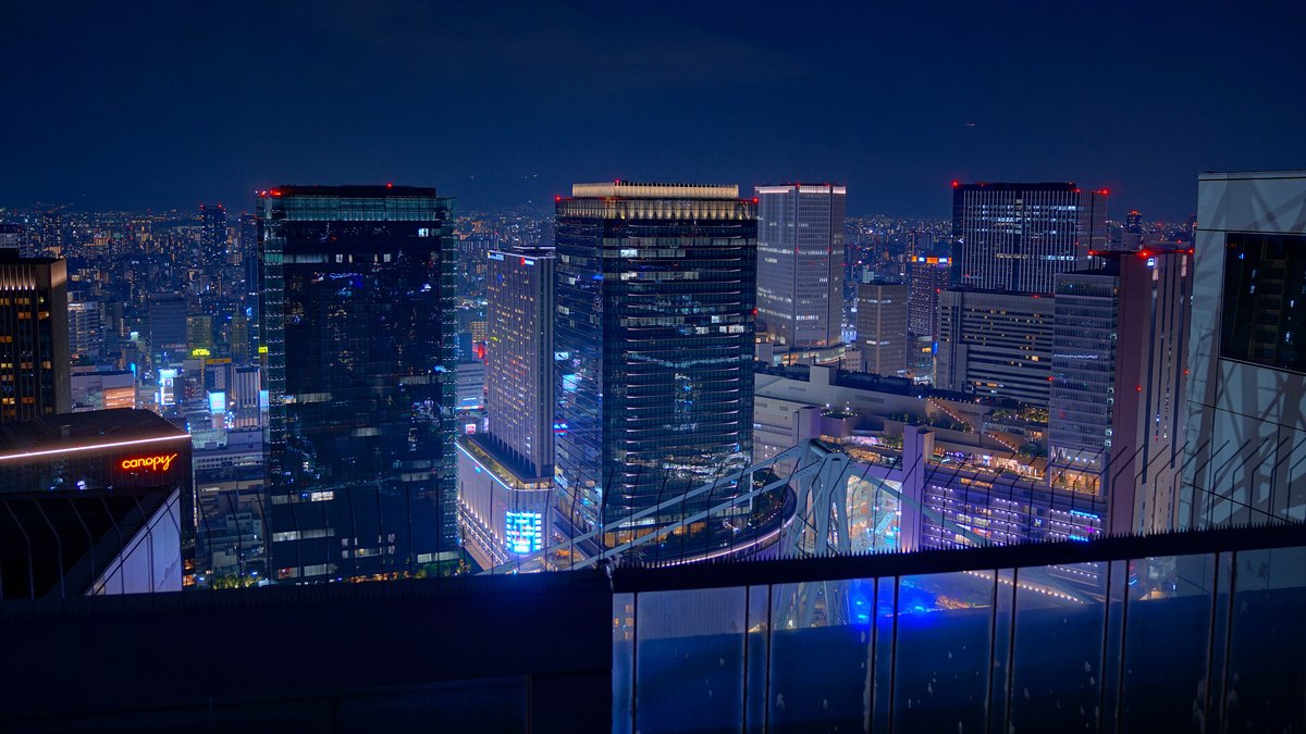 2025 年 8 月夜晚從梅田藍天大廈展望台往東看:前景是展望台的玻璃圍欄,下方看得到連接兩棟樓的圓環結構一角,遠處是大阪站一帶亮著燈的幾棟高樓與整片市區夜景
