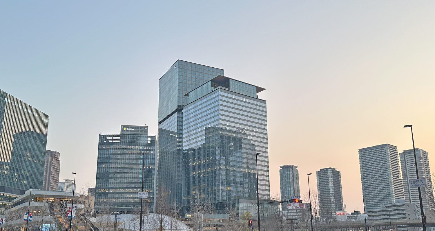 The Umekita area north of Osaka Station at dusk: a glass tower capped by a projecting roof canopy stands at the centre, office blocks and residential towers to either side, street trees and signal posts in the foreground, and a white street-name plate reading Umekita Chuo on a lamp post at the right, under a sky grading from grey-blue to orange