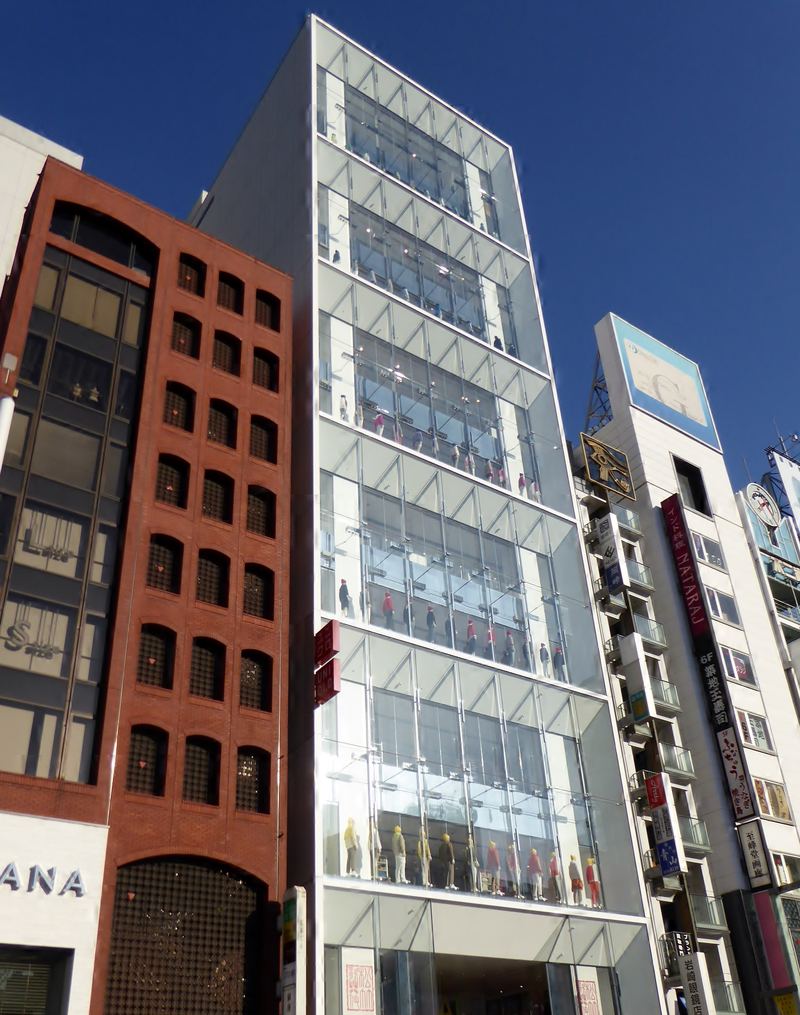 銀座街邊一棟整面玻璃帷幕的高樓，每一層玻璃後面都站著一排穿彩色衣服的人形模特兒，大樓左側外牆掛著兩塊紅色的 UNIQLO 小招牌，兩旁是紅磚大樓與掛滿招牌的白色大樓，天空晴朗