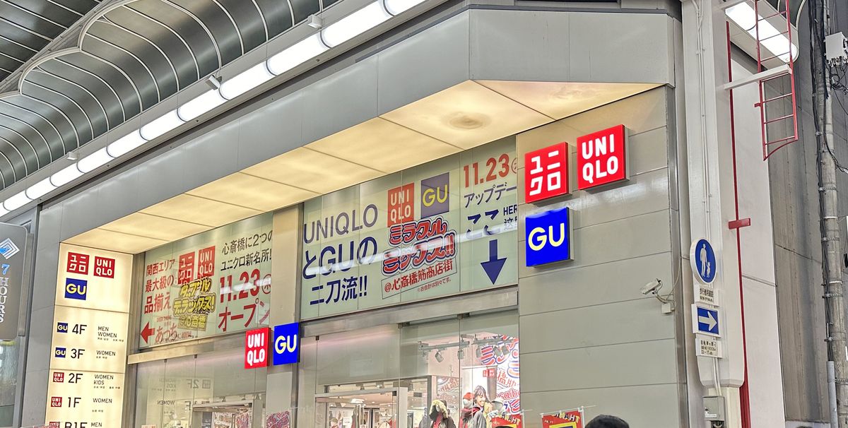 心齋橋筋商店街拱廊下的一棟店面：外牆上紅色的 UNIQLO 與藍色的 GU 方形招牌上下並排，玻璃牆貼著「UNIQLO と GU の二刀流!!」與「11.23 オープン」的大字，左側樓層牌標出 GU 在 3、4 樓、UNIQLO 在 2 樓以下