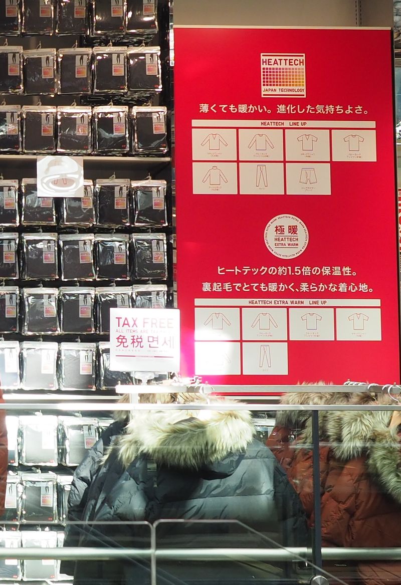UNIQLO 賣場的一面牆：左邊一格一格掛滿袋裝的 HEATTECH 發熱衣，右邊是紅色說明板，上半寫 HEATTECH、下半寫極暖 HEATTECH EXTRA WARM 並畫出各款式的線稿，板子左下夾著一張寫 TAX FREE、免税的白色小牌，前方掛著兩件毛領羽絨外套