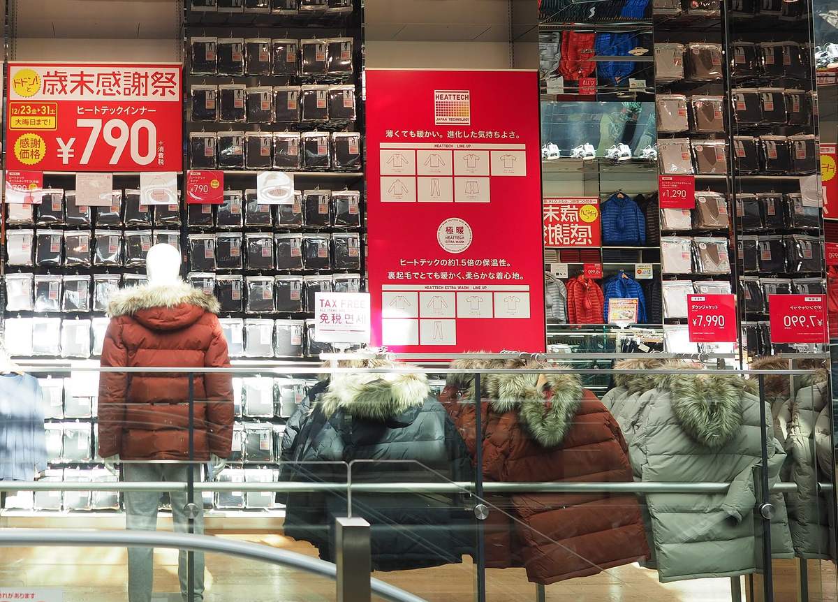 UNIQLO 賣場裡整面牆的袋裝 HEATTECH 發熱衣，中間是紅色的 HEATTECH 與極暖說明板，層架上夾著一張寫 TAX FREE、免税的小牌，前方是一排毛領羽絨外套與穿著外套的人形模特兒
