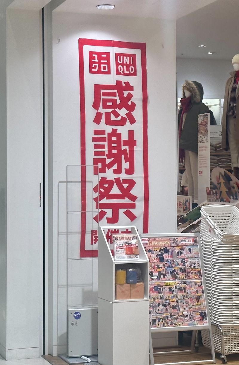 商場裡的 UNIQLO 門市入口：白牆上掛著一幅紅框白底的直式布條，印著 UNIQLO 標誌與紅色大字「感謝祭 開催中」，布條前面是一座擺著馬克杯的白色展示架、一塊貼滿傳單的立牌與一疊白色購物籃，店內的人形模特兒穿著毛領外套與大衣
