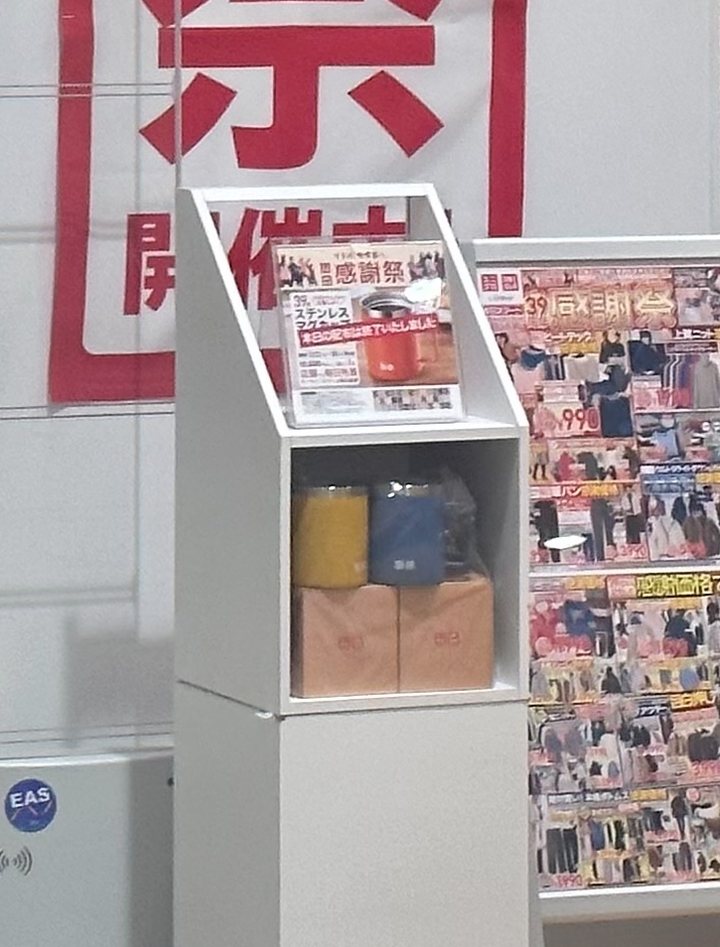 UNIQLO 門市入口的白色展示架：上層斜面放著一張印有「感謝祭」「ステンレス」字樣與橘色馬克杯照片的說明卡，卡片上斜貼一條紅底白字的字條，下層擺著黃色與深藍色兩個馬克杯和兩個紙盒，後面是紅字布條的下半，右邊是貼滿傳單的立牌