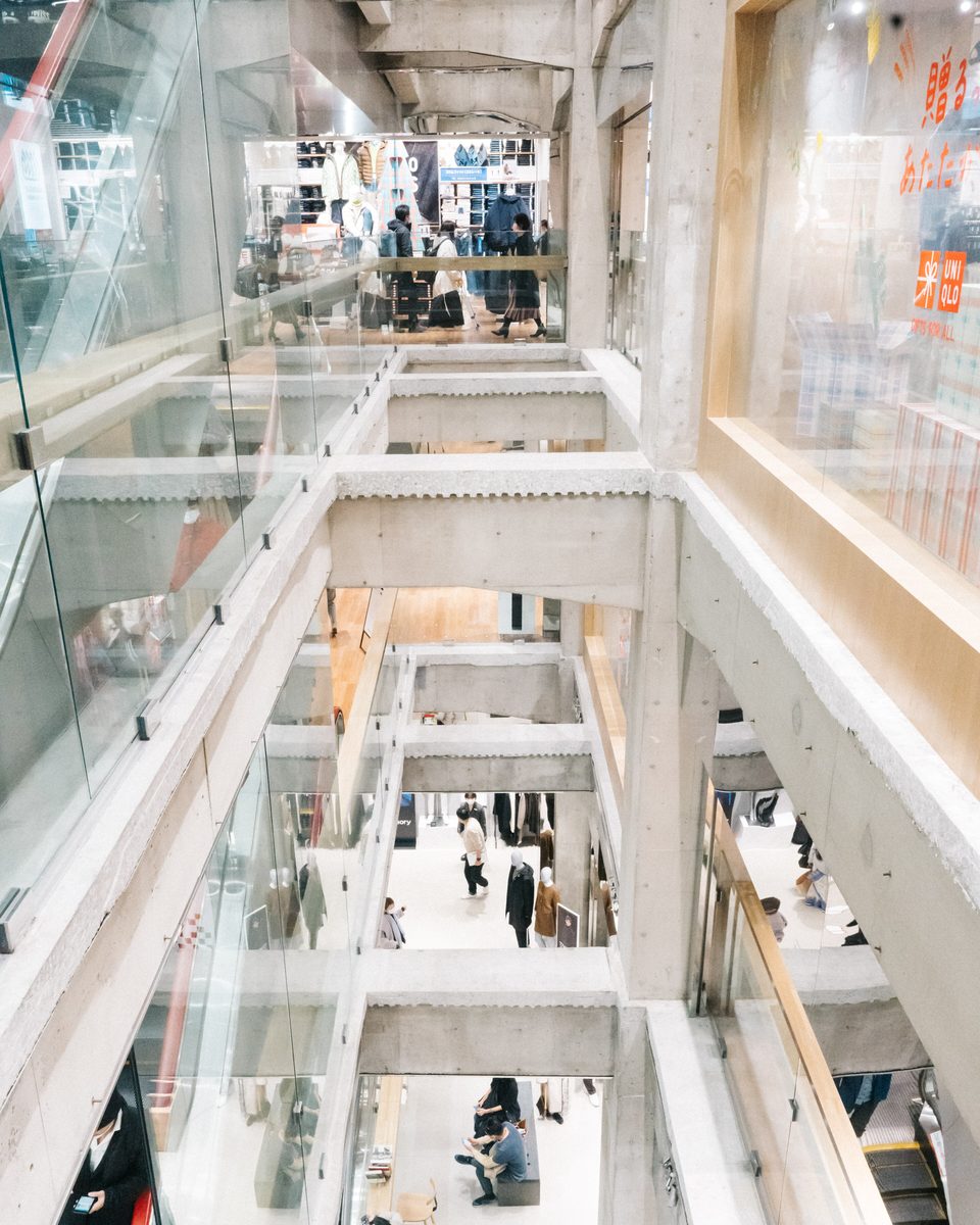 從高樓層往下看 UNIQLO TOKYO 的挑高中庭：清水混凝土橫樑一層層交錯，上方樓層的玻璃欄杆後面是整面牛仔褲層架與穿外套的人形模特兒，下面幾層有逛街與坐著休息的人