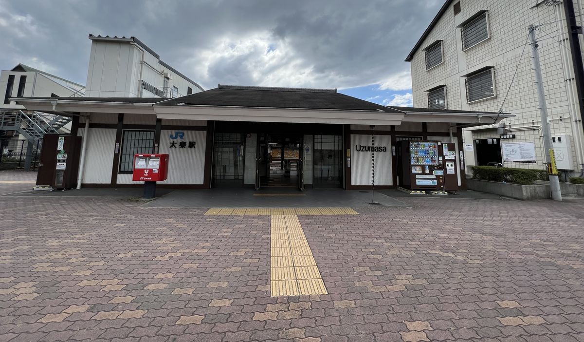 JR 太秦站的站房:平房式的黑瓦屋頂,入口左邊寫「JR 太秦駅」,右邊寫「Uzumasa」,門前有紅色郵筒與自動販賣機,前面是空曠的地磚廣場與導盲磚