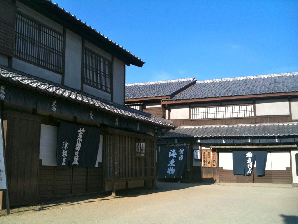 外景區的江戶街道一角:左邊是二樓有格子窗的木造町家,掛著寫「荒物屋」「津軽屋」的深藍布簾,右後方是瓦頂長屋,布簾寫著「諸國 海産物」「物産問屋」,前面是空曠的碎石路,藍天